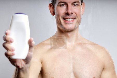 Téléchargez les photos : Close-up portrait of handsome guy showing bottle of gel or shampoo, for advertisement, isolated on gray studio background. young male of caucasian appearance smiling, pleasant and nice, shirtless - en image libre de droit