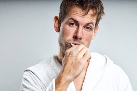 Téléchargez les photos : Confused guy brushing tooth in early morning, after waking up. isolated on gray. caucasian man in white bathrobe look at camera with frustrated facial expression, having no enough sleep - en image libre de droit