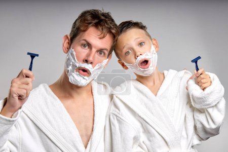 Téléchargez les photos : Caucasian male and little boy having fun with shaving foam on faces in bathroom, isolated. Funny Happy Father and son shaving faces in the morning together, making crazy faces, weekends - en image libre de droit