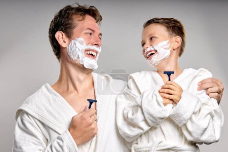Téléchargez les photos : Happy dad and son with pleasant appearance, have shaving foam on faces, hold razors and going to shave, stand together, have fun, laughing. caucasian or american kid imitates father, isolated on gray - en image libre de droit