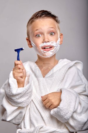 Téléchargez les photos : Surprised child boy son with pleasant appearance, have shaving foam on face, hold razor and going to shave, stand in frot of mirror isolated on gray background, young kid imitates father, in bathrobe - en image libre de droit