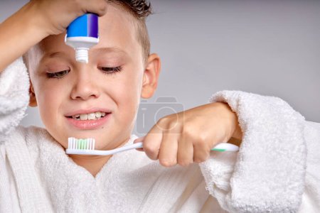 Téléchargez les photos : Child going to brush teeth. Kid applying paste on tooth brush, wearing bathrobe in the morning. Dental hygiene and health for children. isolated gray studio background, close-up portrait - en image libre de droit
