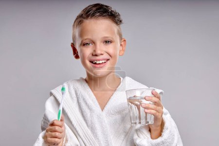 Téléchargez les photos : Smiling child holding teeth brush and glass of water in hands, Dental hygiene and health for children. plasant kid in white bathrobe enjoy morning routine, happy, isolated on gray background - en image libre de droit
