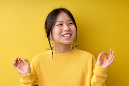 Téléchargez les photos : Funny cheerful asian woman meditating over yellow isolated background relax and smiling, looking at side, doing meditation gesture with fingers. Yoga concept. Portrait, copy space - en image libre de droit