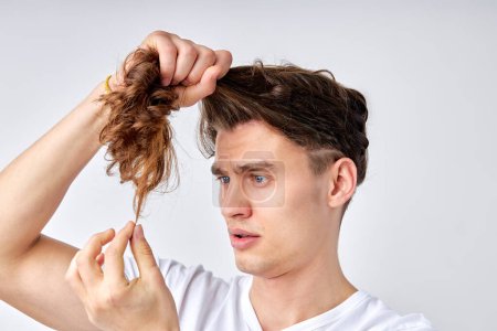Foto de Amazed confused male looking at split hair ends, want to cut isolated over light gray background. european young guy model in casual wear looking embarrassed, need hair care. portrait copy space - Imagen libre de derechos