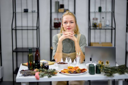 Photo for Bottles with fragrance on table with many bottles of another essential oils are used for testing scent by female perfumer. portrait of confident blonde lady in apron uniform, using laptop during work - Royalty Free Image