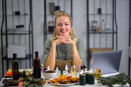 Foto de Bottles with fragrance on table with many bottles of another essential oils are used for testing scent by female perfumer. portrait of pleasant blonde lady in apron uniform, using laptop during work - Imagen libre de derechos