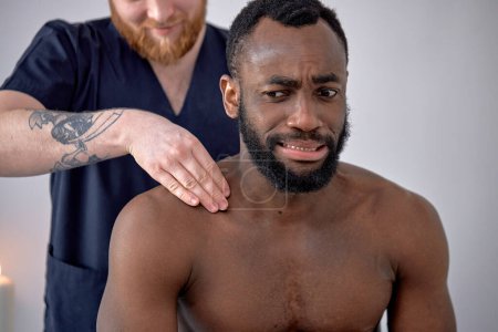 Photo for Cropped caucasian Spa therapist making relaxing massage for handsome middle aged black man, rubbing back. Frustrated bearded man getting healing spine massage at modern luxury spa - Royalty Free Image