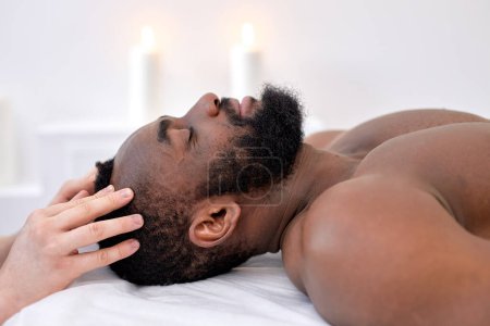 Téléchargez les photos : Head massage in spa wellness salon. recovery treatment. Wellness. confident black guy client lying on couch bed, relaxing, with eyes closed. side view. healthy lifestyle, therapy - en image libre de droit
