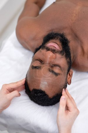 Foto de Spa therapist making relaxing massage for handsome middle aged black man. Peaceful bearded man getting healing head massage at modern luxury spa. handsome guy lying on bed couch relaxed, top view - Imagen libre de derechos