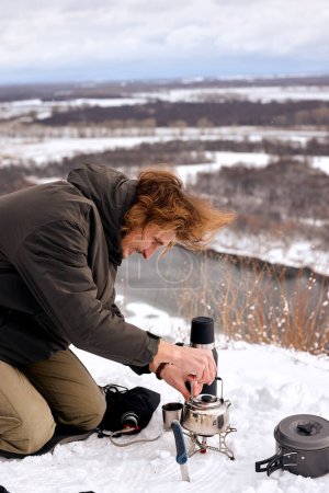 Photo for Male put small tourist kettle on fire in winter forest during camping, travelling. alone. river or lake in the background, wonderful landscape. travel, camping, adventure concept - Royalty Free Image