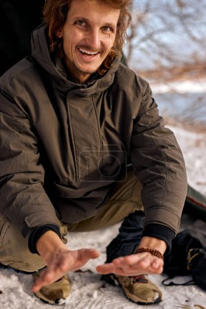 Foto de Smiling man warm hands by fire, sit next to fire. Travel concept. happy guy enjoy exploring new places, camping. Burning wood at evening in nature, in winter forest, alone. portrait - Imagen libre de derechos