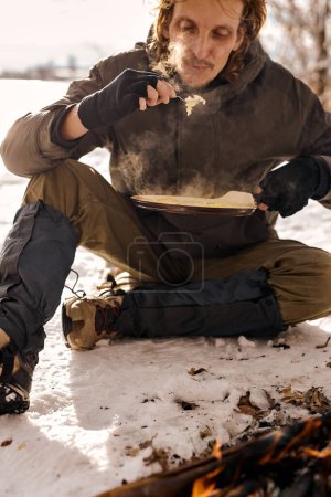 Photo for Young caucasian man traveler cook scrambled eggs on fire. bushcraft, adventure, travel, tourism, hike and camping concept. handsome guy in warm coat sit going to eat delicious food on nature - Royalty Free Image