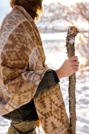 Téléchargez les photos : Young male tourist with wooden stick looking around while walking in winter forest on cold day, wrapped in scarf, at sunny frozen day or morning, bushcraft, travel, nature, active lifestyle concept - en image libre de droit