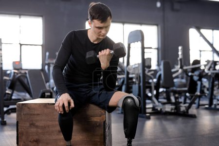 Photo for A young strong handsome motivated man with a disability with a prosthetic leg is training in a gym with dumbbells. Motivation poster. copy space, lifestyle, free time, spare time - Royalty Free Image