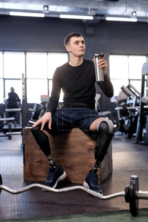 Photo for Sweaty bodybuilder with thermos in hand takes break after workout with barbell looks sideways at gym, full length shot. take a break. - Royalty Free Image