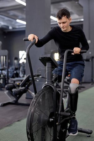 Foto de Hombre fuerte haciendo ejercicio físico para la recuperación de lesiones hombre usando bicicleta estacionaria para aeróbicos y gimnasia para tratar el dolor muscular - Imagen libre de derechos