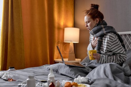 Foto de Enferma mujer caucásica tiene mala tos, sentado en la cama en casa, beber té caliente y trabajar en el ordenador portátil. Mujer joven con fuerte dolor de cabeza. covid-19, coronavirus, concepto omicrón - Imagen libre de derechos