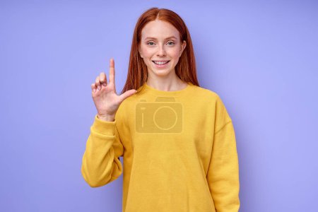 Foto de Hermosa mujer pelirroja alegre alegre en suéter elegante amarillo demostrando letra L símbolo de lenguaje de signos para sordos humanos con fondo azul. retrato de primer plano aislado - Imagen libre de derechos