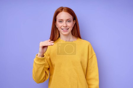 Foto de Hermosa mujer pelirroja feliz en suéter elegante amarillo demostrando letra T símbolo de lenguaje de signos para sordos humanos con fondo azul. retrato de primer plano aislado - Imagen libre de derechos