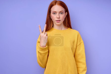 Téléchargez les photos : Belle femme rousse sérieuse confiante en pull élégant jaune démontrant le symbole de langue des signes de la lettre V pour les sourds humains avec un fond bleu. portrait isolé en gros plan - en image libre de droit
