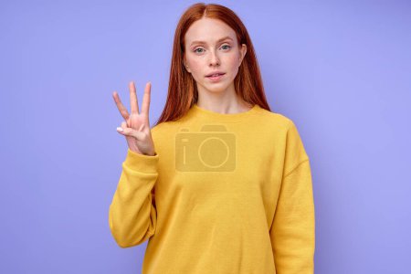 Foto de Hermosa mujer pelirroja de confianza seria en suéter elegante amarillo demostrando letra W símbolo de lenguaje de signos para sordos humanos con fondo azul. retrato de primer plano aislado - Imagen libre de derechos