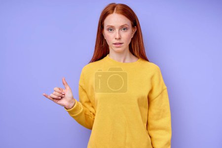Foto de Hermosa mujer pelirroja de confianza seria en suéter elegante amarillo que demuestra la letra Y símbolo de lenguaje de signos para sordos humanos con fondo azul. retrato de primer plano aislado - Imagen libre de derechos