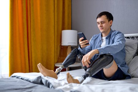 Photo for Man with artificial leg prosthesis using smartphone, browsing web or watching movie at home. Handicapped young Caucasian guy checking social media, typing message to friend on mobile device - Royalty Free Image