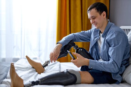 Photo for Cheerful disabled male with artificial leg stay at home. adult guy with prostheses on legs having rest. young Caucasian man in casual wear sit on bed using smartphone, typing message - Royalty Free Image