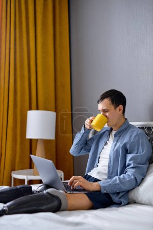 Photo for Young man with prosthesis on legs using laptop for remote job at home, sitting on bed and drinking tea. Confident Caucasian guy with physical disability having online work. never give up concept - Royalty Free Image