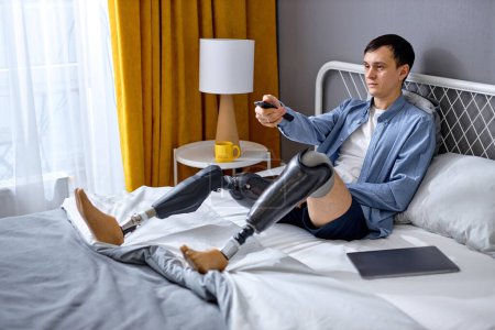 Photo for Lonely disabled Caucasian guy with TV controller enjoying movie indoors. Nice young handicapped man with prosthesis watching television, switching between channels, alone at home, in casual wear - Royalty Free Image