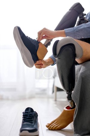 Foto de Hombre discapacitado irreconocible amputado pierna usando zapatillas de deporte en la prótesis en casa sentado en la cama, dificultades para discapacitados después de lesiones por accidente. tipo preparándose para el día - Imagen libre de derechos