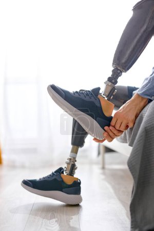 Téléchargez les photos : Recadré homme handicapé non reconnaissable jambe amputé portant des baskets sur la prothèse à la maison assis sur le lit, difficultés handicapées après des blessures accidentelles. gars se préparant pour la journée - en image libre de droit