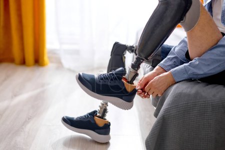 Téléchargez les photos : Réhabilitation après amputation des jambes, homme s'habituant à la prothèse sur les jambes, portant des baskets assises sur le lit dans une chambre lumineuse à la maison. difficultés dans la vie des personnes handicapées, handicap physique - en image libre de droit