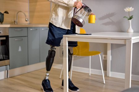 Téléchargez les photos : Handicapé mâle plomb mode de vie habituel comme d'autres personnes, se tenir dans artificielles fausses jambes passer la routine du matin. handicapé homme méconnaissable avec prothèse de jambe dans la cuisine à la maison, faire du thé - en image libre de droit
