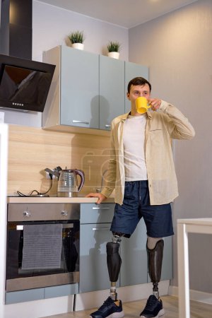 Foto de Pie masculino en piernas falsas artificiales beber café o té en la cocina, pasar la rutina de la mañana. hombre caucásico discapacitado con prótesis disfrutar de la mañana o el día. persona discapacitada feliz - Imagen libre de derechos