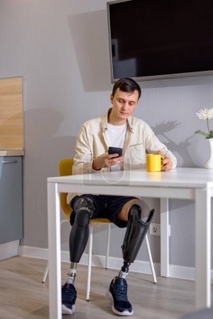 Foto de Relajado chico apuesto discapacitado con biónico piernas inteligentes prótesis sentarse en la cocina, viendo la película en el teléfono inteligente y beber café o té, solo, en el interior. discapacidad física, concepto de personas - Imagen libre de derechos
