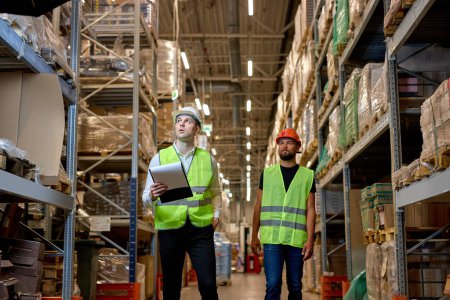 Photo for Retail Warehouse full of Shelves with Goods, Two Caucasian Male Workers Supervisors in Working Uniform and Helmet Discuss Product Delivery. Workers Walking and Having Talk In Warehouse - Royalty Free Image