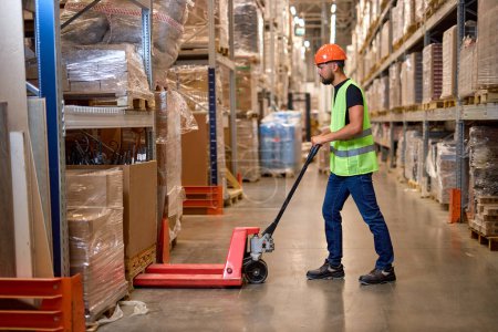 Photo for Young male warehouse worker carrying delivery to production stock using forklift. male staff in vest and orange helmet in place of work. Forklift driver stacking pallets with packs - Royalty Free Image