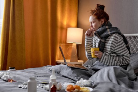 Foto de Mujer caucásica enferma tiene mala tos, sentado en la cama en casa, beber té caliente y trabajar en línea en el ordenador portátil desde casa. Mujer joven con fuerte dolor de cabeza. covid-19, coronavirus, concepto omicrón - Imagen libre de derechos