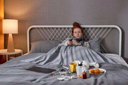 Foto de Mujer de jengibre pálido enfermo acostado en la cama y con control remoto de televisión, encendiendo, apagando la televisión, televisión retrato de cerca - Imagen libre de derechos