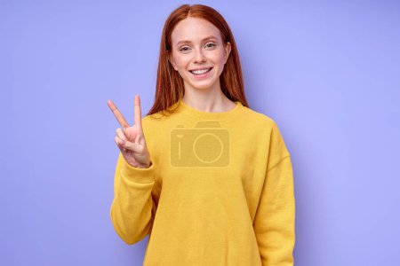 Photo for Young smiling satisfied cheerful red-haired Caucasian woman looking at camera showing victory gesture isolated on blue background studio shot closeup portrait, happiness satisfaction body language - Royalty Free Image