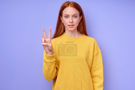 Photo for Beautiful happy cheerful redhead woman in yellow stylish sweater demonstrating letter N sign language symbol for deaf human with blue background. isolated close up portrait - Royalty Free Image