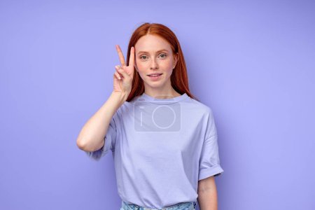 Photo for Attractive redhead girl showing two fingers looking at camera, victory sign. close up portrait isolated blue background. woman demonstrating the letter V. - Royalty Free Image