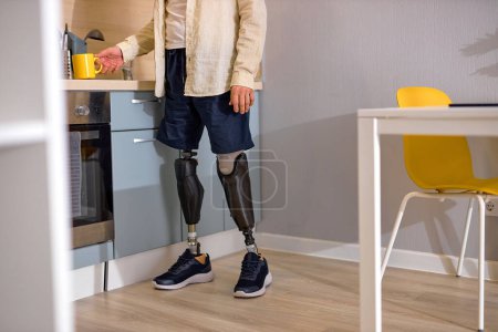 Photo for Cropped handicapped or disabled person with disability standing on bionic smart prosthesis in kitchen, making coffee or tea for himself at home. indoors. in the morning. disability lifestyle - Royalty Free Image
