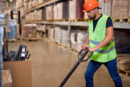 Photo for Male staff worker in warehouse uses hand pallet stacker to transport goods, alone, dressed in working clothes and safety hard hat.Skilled warehouse employee pushing manual pallet jack - Royalty Free Image