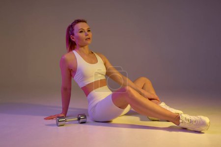 Photo for Fitness young tired sportswoman in white top and leggings taking break between exercises at home. Caucasian fair haired woman sitting on floor near sport dumbbells. full length brown background - Royalty Free Image