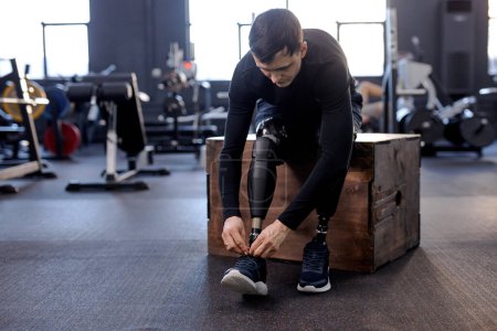 Photo for Amputee male athlete tying shoelaces sitting on fitness box in gym with modern equipment, full length photo. health and body care - Royalty Free Image