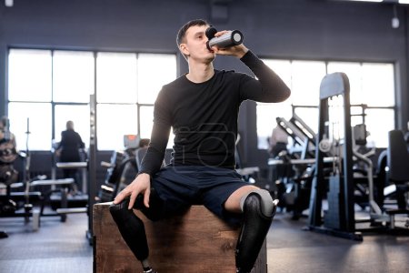 Photo for A thirsty tired handsome sportsman with artificial leg sitting at sport center drinking refreshment. close up photo, free time, spare time, lifestyle - Royalty Free Image