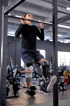 Foto de Hombre ambicioso activo con la pierna protésica que usa ropa deportiva que hace pull-ups en la barra en el gimnasio moderno, tiro de vista lateral de longitud completa, tiempo libre, tiempo libre, estilo de vida - Imagen libre de derechos